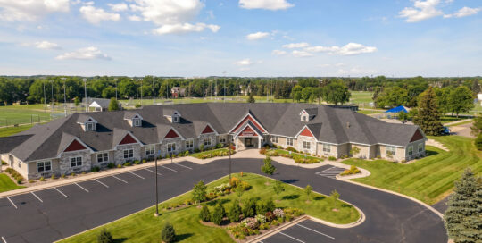 Front exterior photo of Suite Living Senior Care of Plymouth. It shows the parking lot and front entrance from about 200 feet in the air.