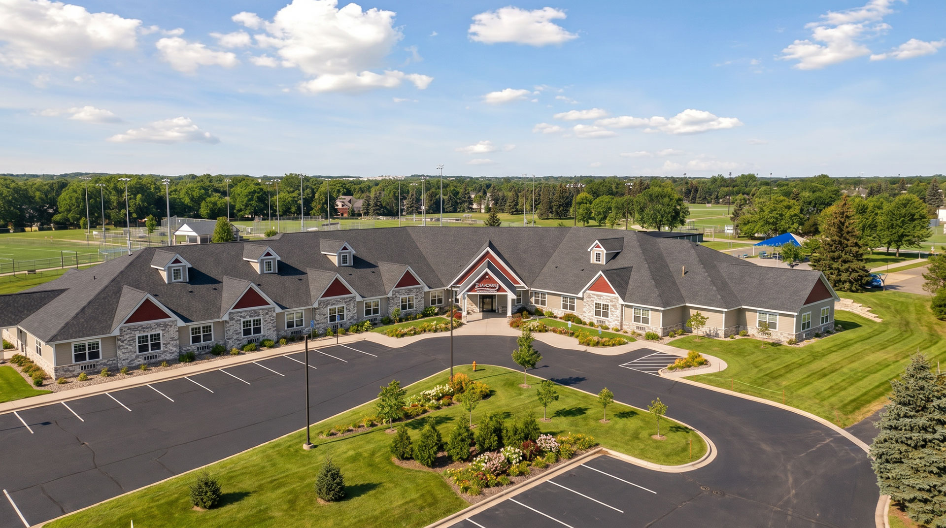 Front exterior photo of Suite Living Senior Care of Plymouth. It shows the parking lot and front entrance from about 200 feet in the air.